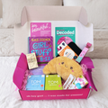 The First Period Box open on a bed with a croissant heat bag, Girl Stuff 8-12 book, Love Luna period undies, TOM Organic pads, drinking chocolate and cards