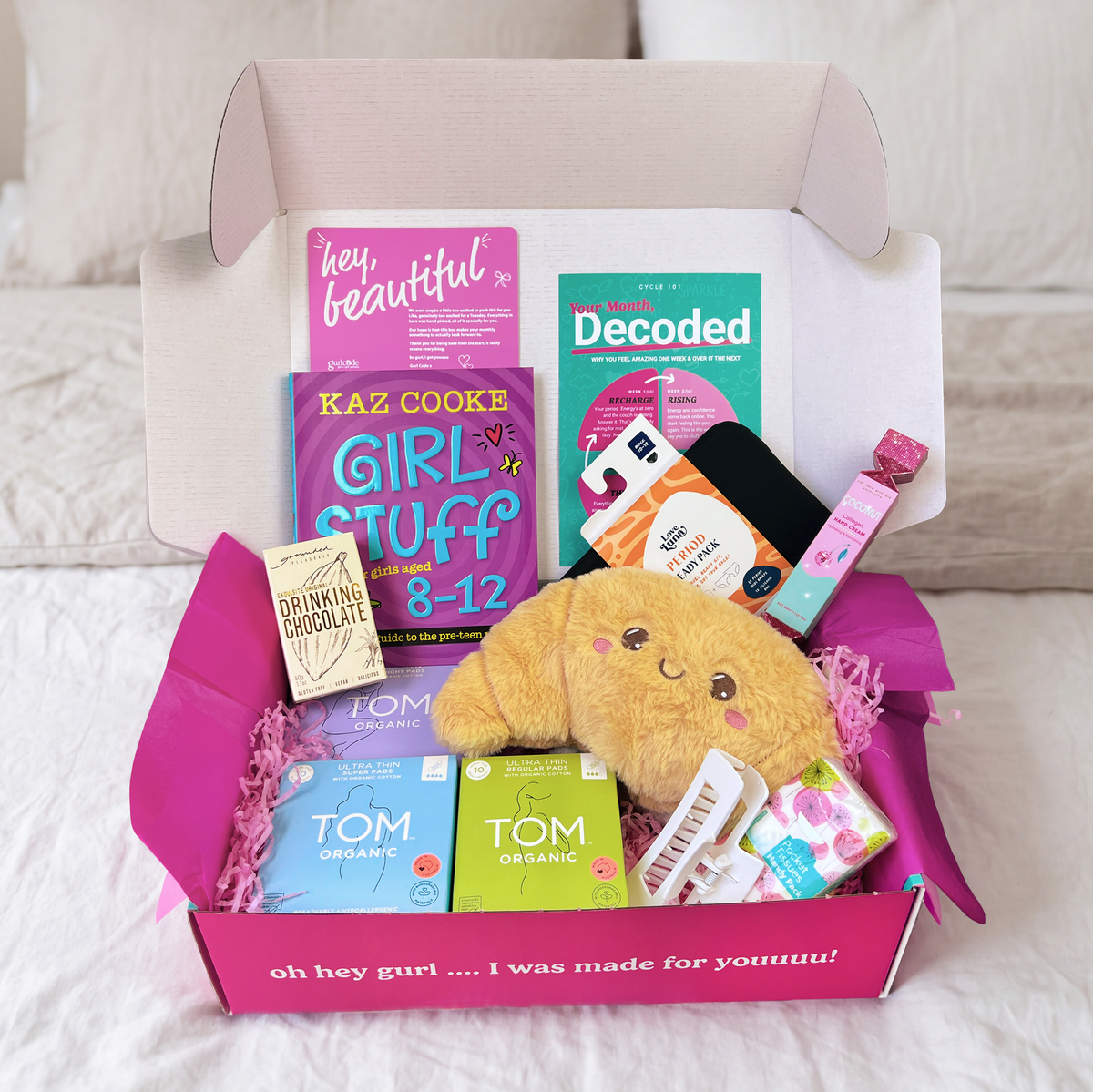 The First Period Box open on a bed with a croissant heat bag, Girl Stuff 8-12 book, Love Luna period undies, TOM Organic pads, drinking chocolate and cards