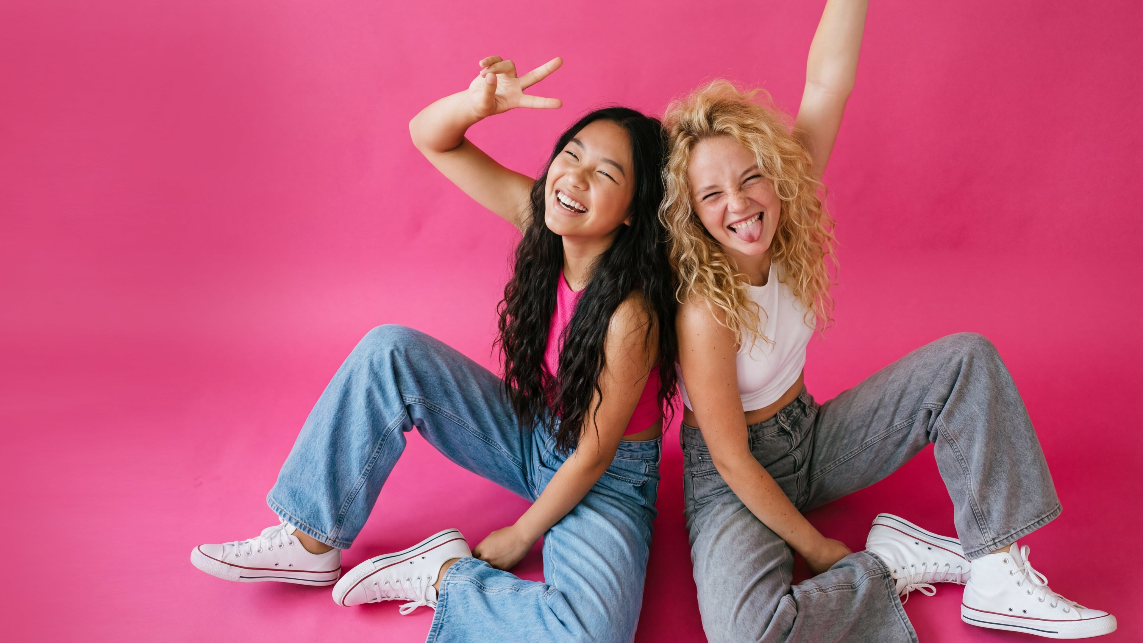 Two people sitting on a pink background, one making a peace sign.
